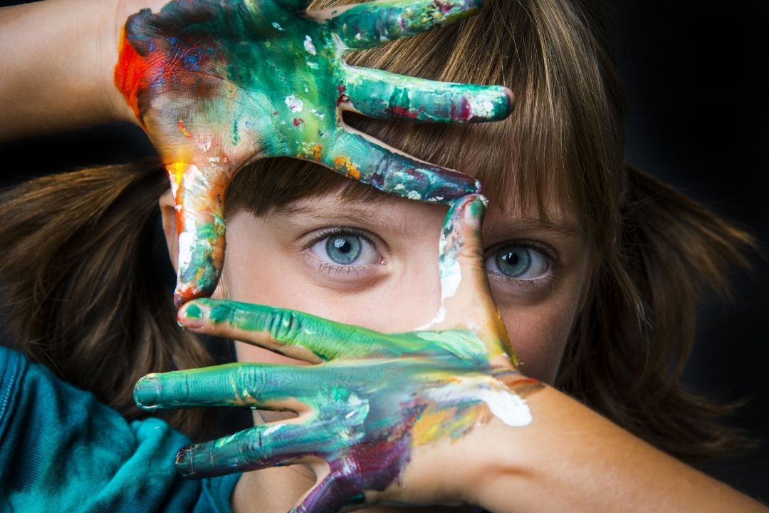 Girl with paint on her hands holds them in front of her face around her eye