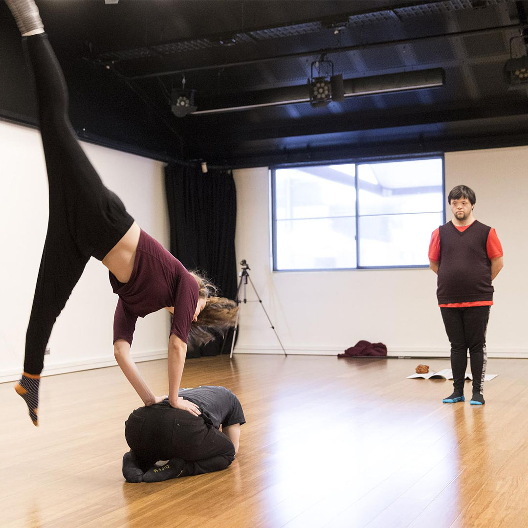 Three dancers in a studio, one is curled up in a ball on the ground with another jumping over them, while the third watches